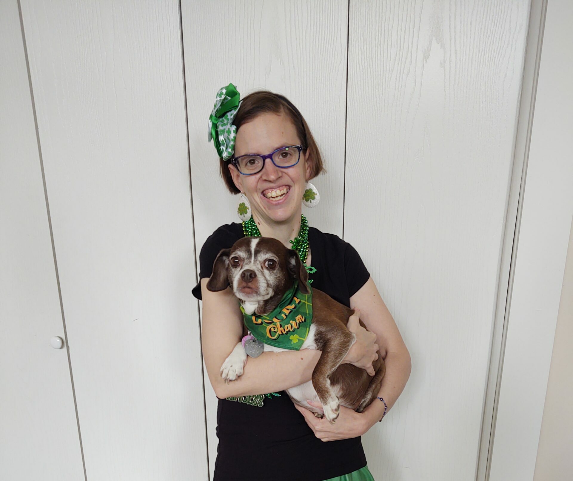 A cheerful woman in glasses, wearing a black shirt and green-themed St. Patrick's Day accessories, holds a brown and white dog wearing a 'Lucky Charm' bandana. They are both in front of a white paneled door, celebrating the festive occasion.