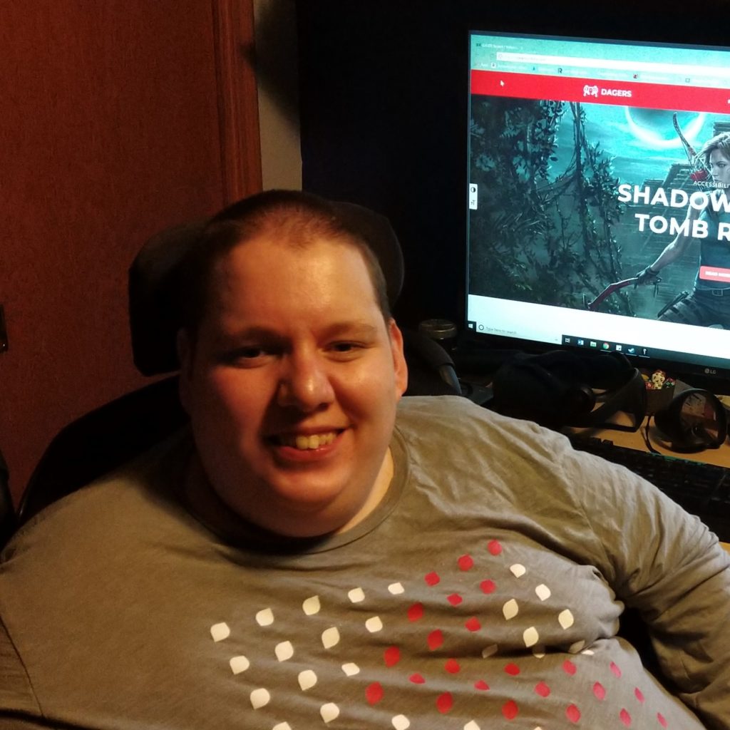 A person with short hair is smiling at the camera, sitting in a wheelchair in front of a computer desk. They wear a grey T-shirt with a pattern of red and white dots. Behind them, a monitor displays a video game title screen, headphones rest on the desk, and various items are scattered around, suggesting a cozy, personal space.