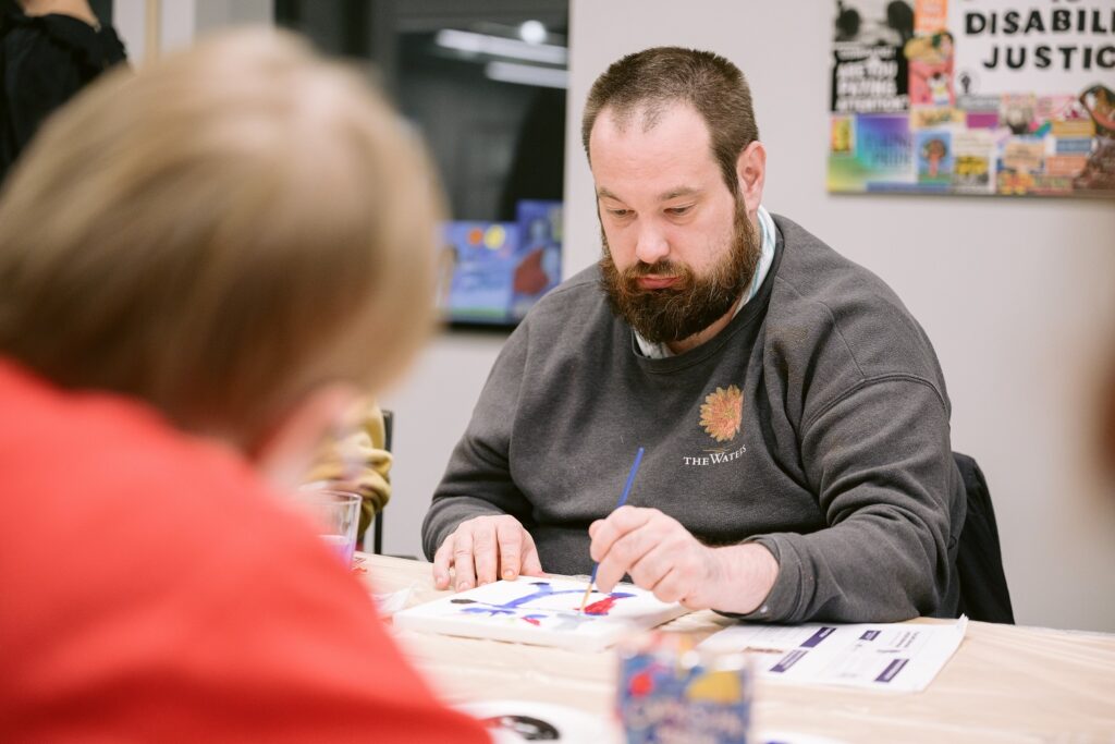 A bearded man in a grey sweatshirt with a leaf motif sits at a table and concentrates on painting with a blue brush on paper, with a woman's profile visible in the foreground and a 'Disability Justice' poster in the background. The man is attending an artistic self-advocacy workshop at The Arc Minnesota.