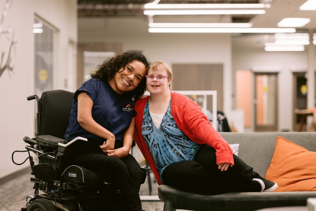 Two happy women posing for a photo, one seated in a power wheelchair wearing a blue t-shirt, the other sitting on a couch wearing glasses and a red cardigan over a blue top. They are in a modern, well-lit indoor space with soft furnishings and office doors in the background