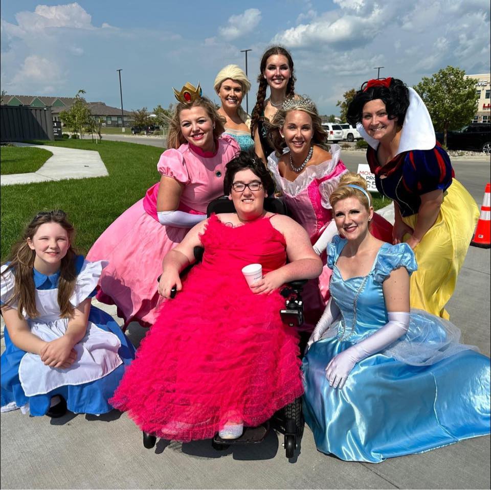 A woman in a wheelchair with short brown hair and glasses is dressed as a Disney princess and is smiling at the camera. There are seven other princesses gathered around her, smiling at the camera.